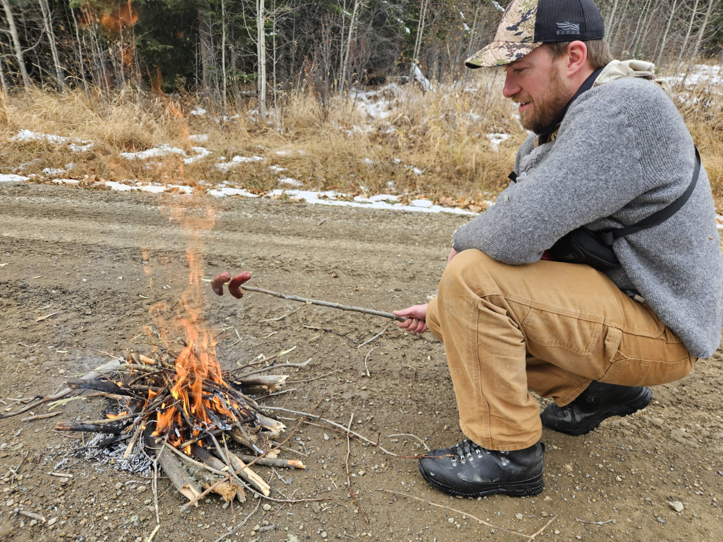 A young male hunter roasts sausages over a cooking fire that has been made on a gravel road, away from fire hazards.