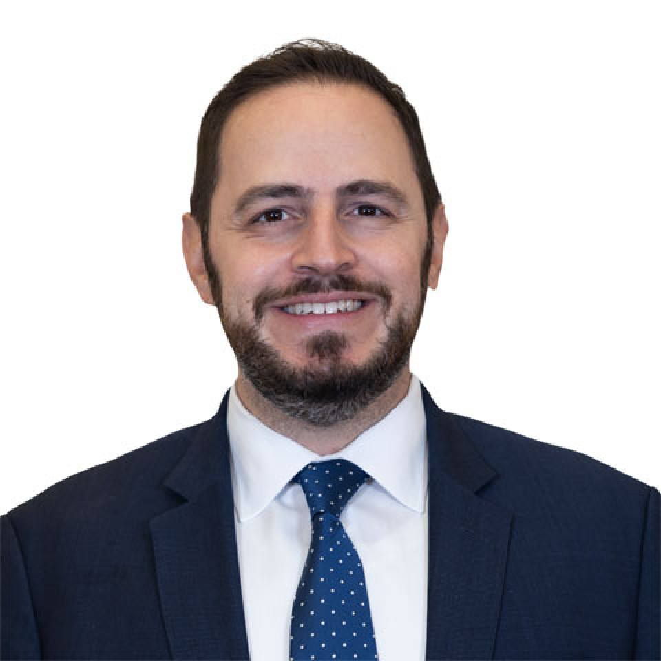 Demetrios Nicolaides is smiling at the camera wearing a dark suit and tie with a white dress shirt.